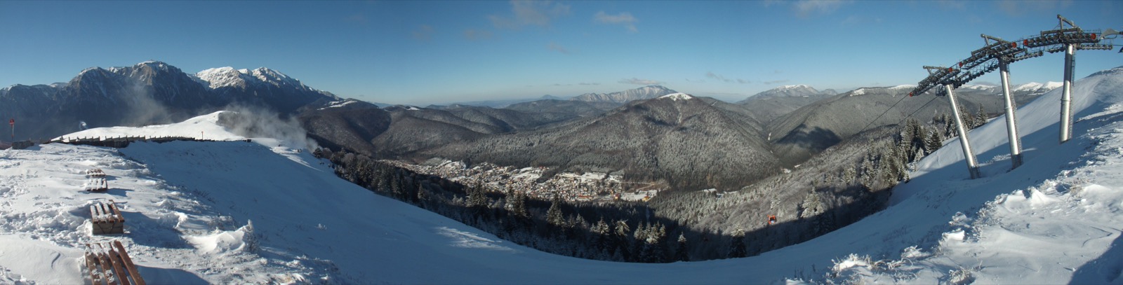 Panoramă de sus, de la capătul pârtiilor: stâlpii telegondolei și o cabină roșie deasupra pădurii, Azuga în vale și Bucegii înzăpeziți în stânga