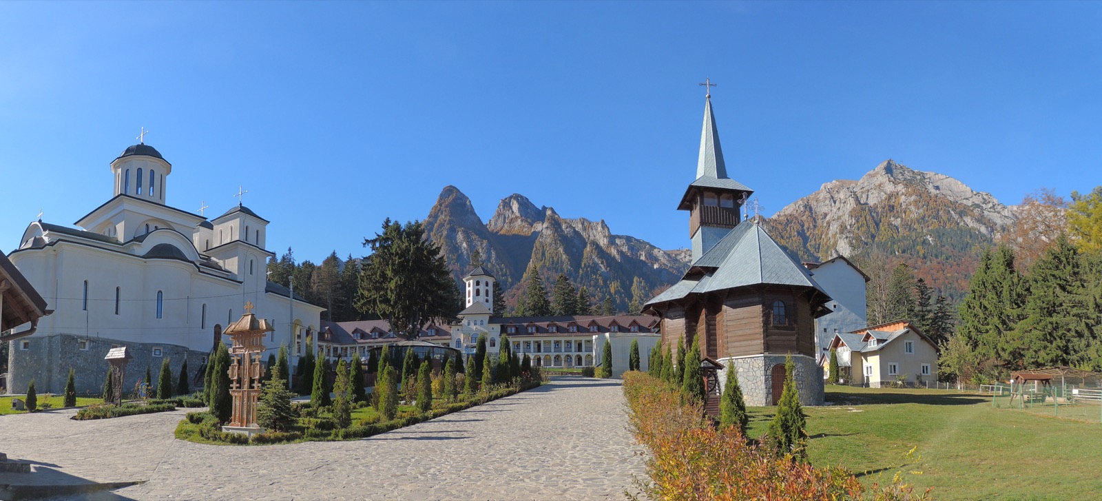 Mănăstirea Caraiman, cu biserica mare de zid, biserica de lemn cu turlă înaltă și crestele Bucegilor în spate
